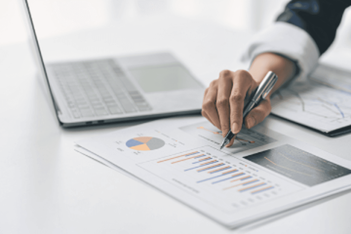 Eine Hand hält einen Stift über Diagramme auf einem weißen Schreibtisch mit einem Laptop. Datenanalyse, Wirtschaft, Finanzen.