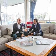 Two men in suits shake hands on a beige couch, with a harbor view through a window. A table holds papers and magazines.