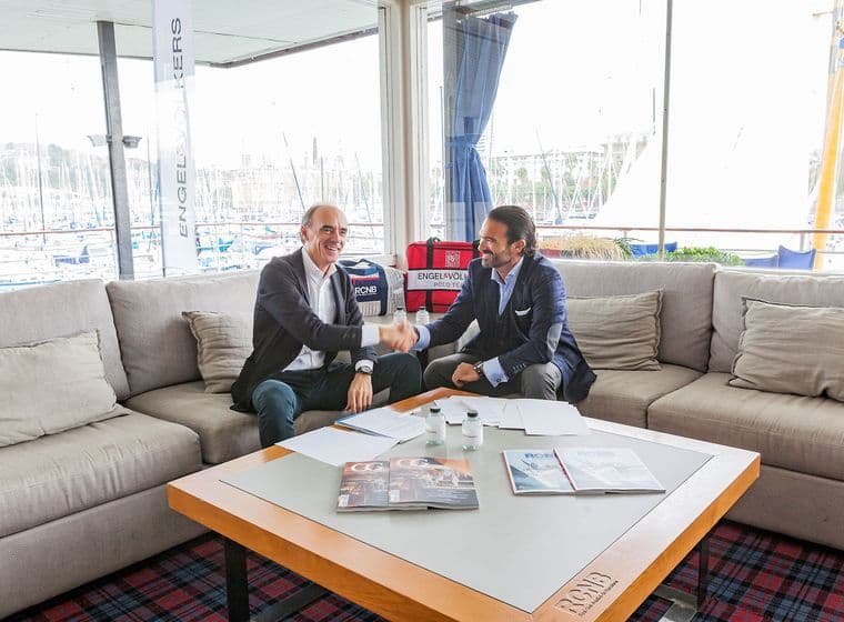 Dos homes en vestit s'estrenyen la mà en un sofà beix, amb una vista del port a través d'una finestra. Una taula sosté papers i revistes.