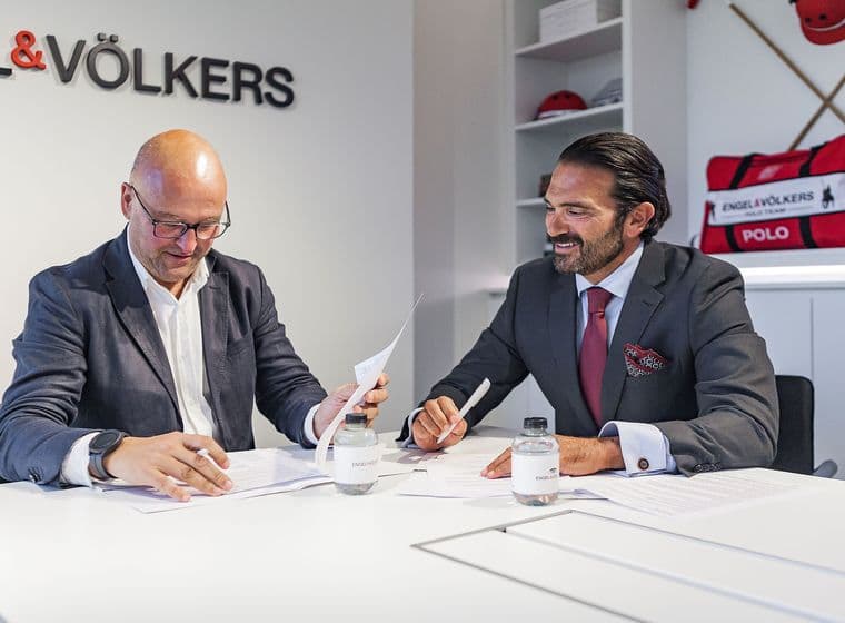 Dos homes en vestit repassen documents en una taula blanca en una oficina d'Engel & Völkers. El logotip de l'empresa és visible a la paret i a les ampolles d'aigua.