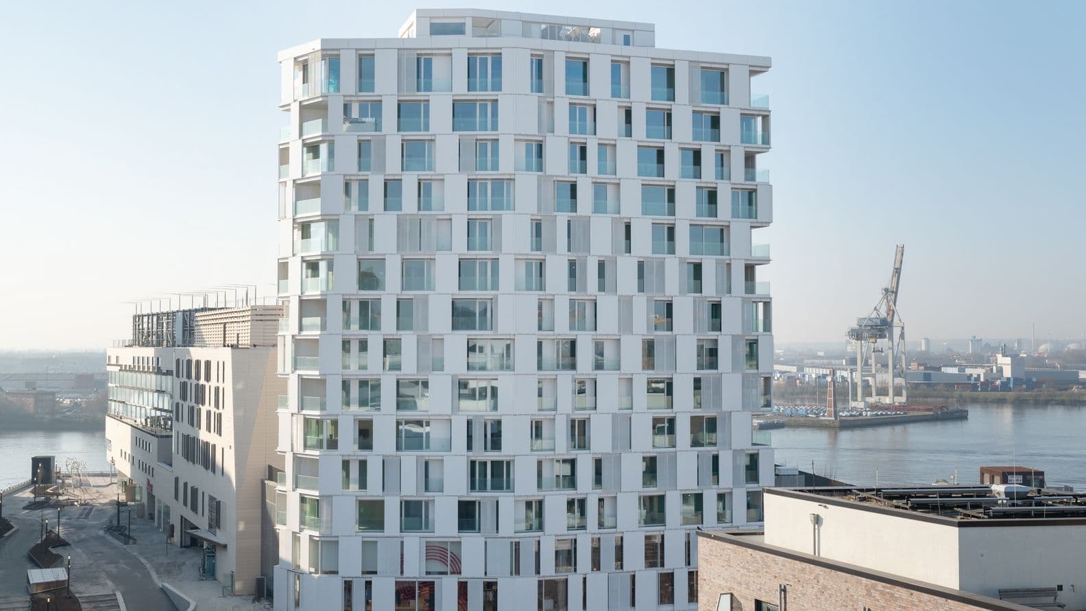 Modern white high-rise building with numerous windows, labeled "The Lyte," near a waterfront with industrial cranes in the background.
