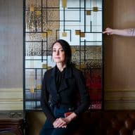 A woman in black sits on a brown leather couch in front of a Mondrian-style glass panel. An arm with a tattoo is holding the panel.