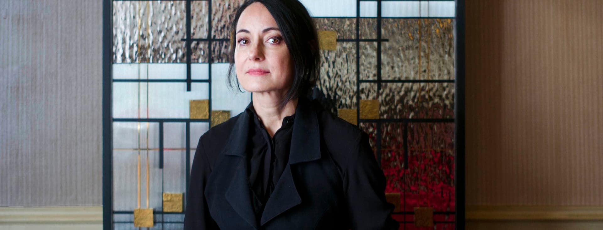 Une femme vêtue de noir est assise sur un canapé en cuir marron devant un panneau de verre de style Mondrian. Un bras avec un tatouage tient le panneau.