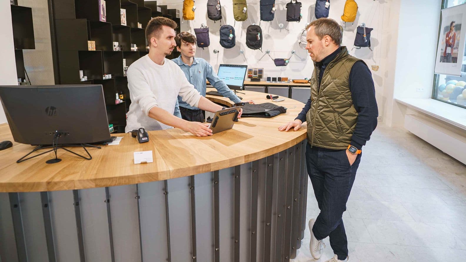 Three men stand by a curved wooden sales counter in a shop, with backpacks displayed on the wall behind them; one man is holding a tablet and speaking with a customer.