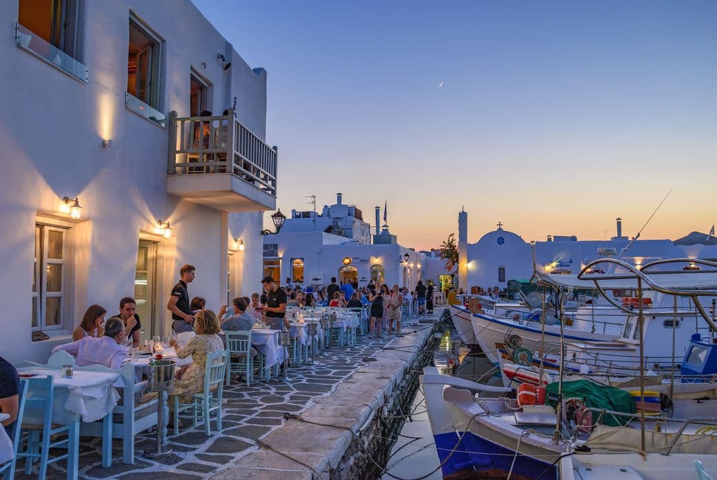 Outdoor restaurant at dusk in Mykonos, Greece. White buildings, tables with diners, and boats in the harbor.