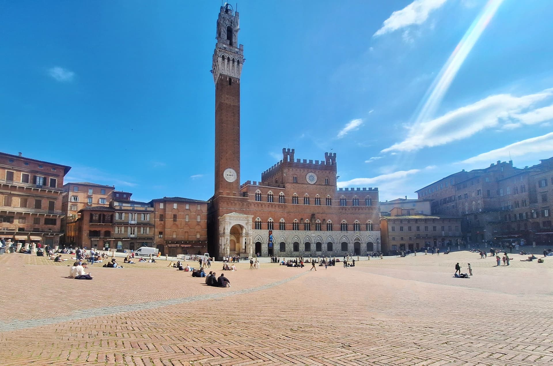 Historic square with a tall brick tower and medieval buildings under a clear blue sky, surrounded by people sitting and walking.
