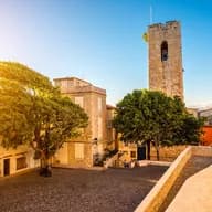 Cobblestone square in Antibes, France, with stone buildings, a tall tower, and a large tree under a clear blue sky.