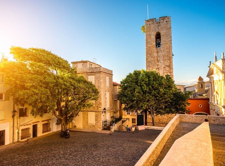 Cobblestone square in Antibes, France, with stone buildings, a tall tower, and a large tree under a clear blue sky.