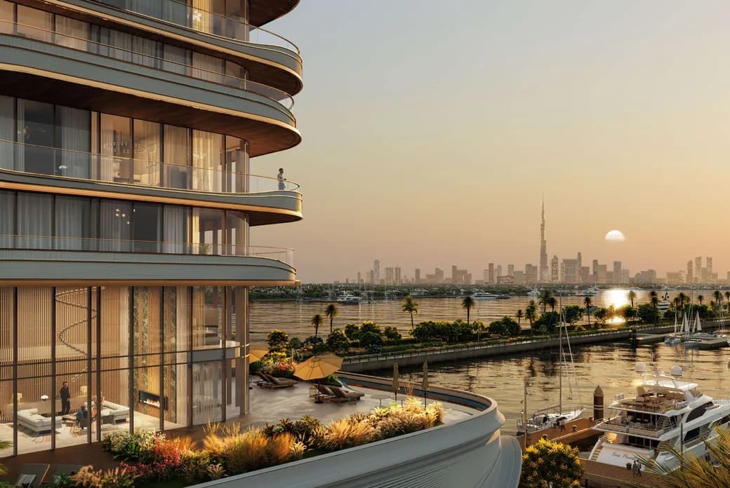 Curved modern building with balconies overlooking a marina and city skyline at sunset, featuring yachts and lush greenery.