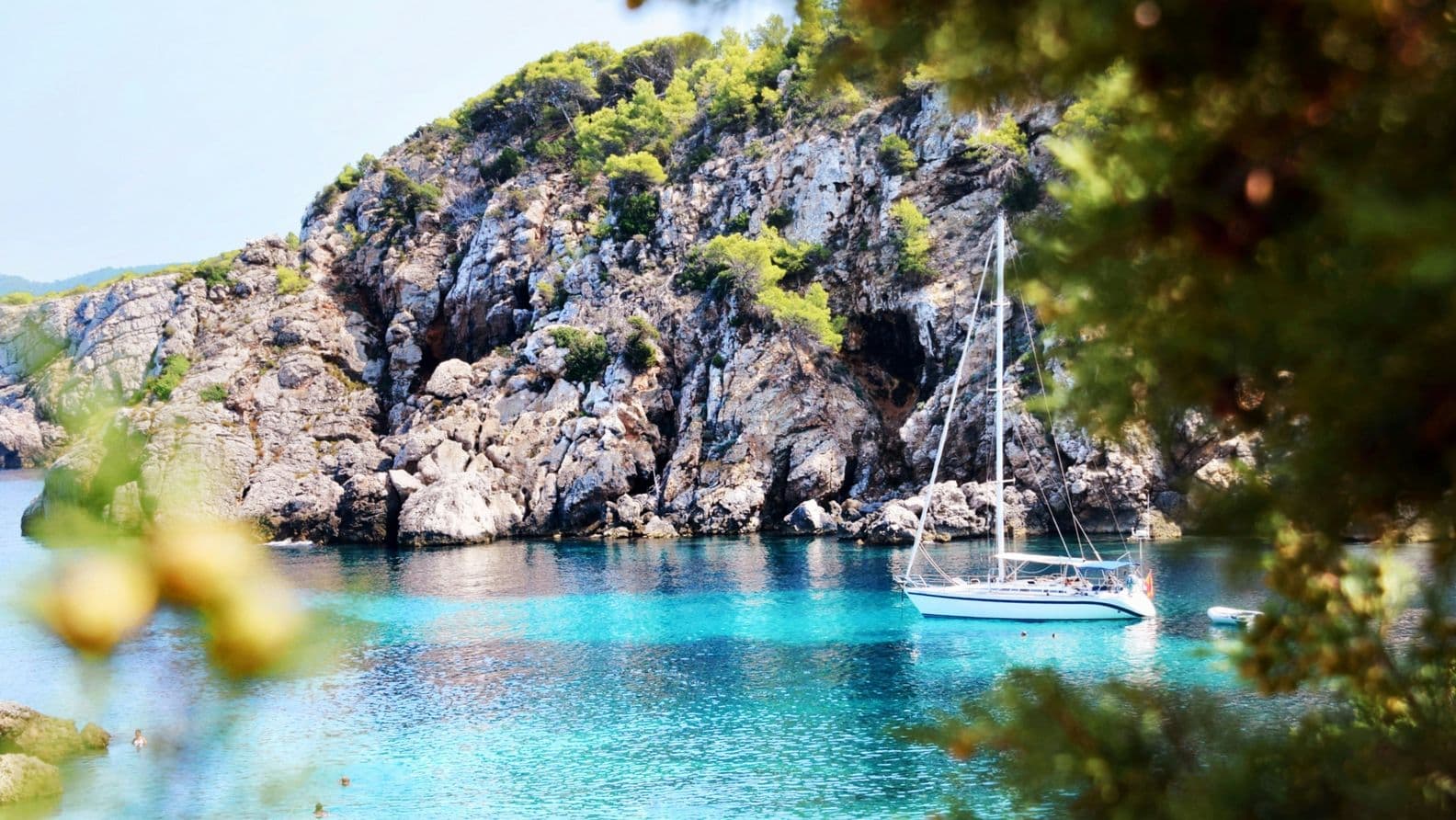 Traumhafte Bucht auf Ibiza mit kristallklarem, türkisfarbenem Wasser und einem elegant dahingleitenden Segelboot – umgeben von felsigen Ufern und der ruhigen Schönheit der Mittelmeerküste.