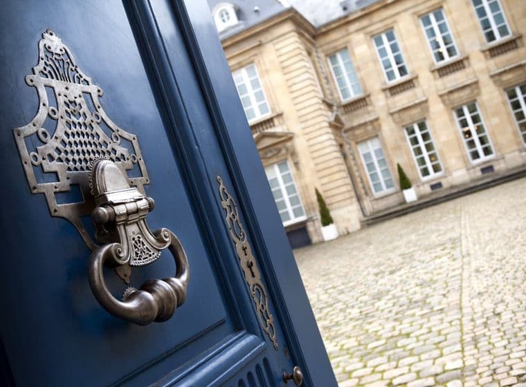 Ajar blue door with ornate metal handle and keyhole, revealing a cobblestone courtyard and a tan building with white windows.