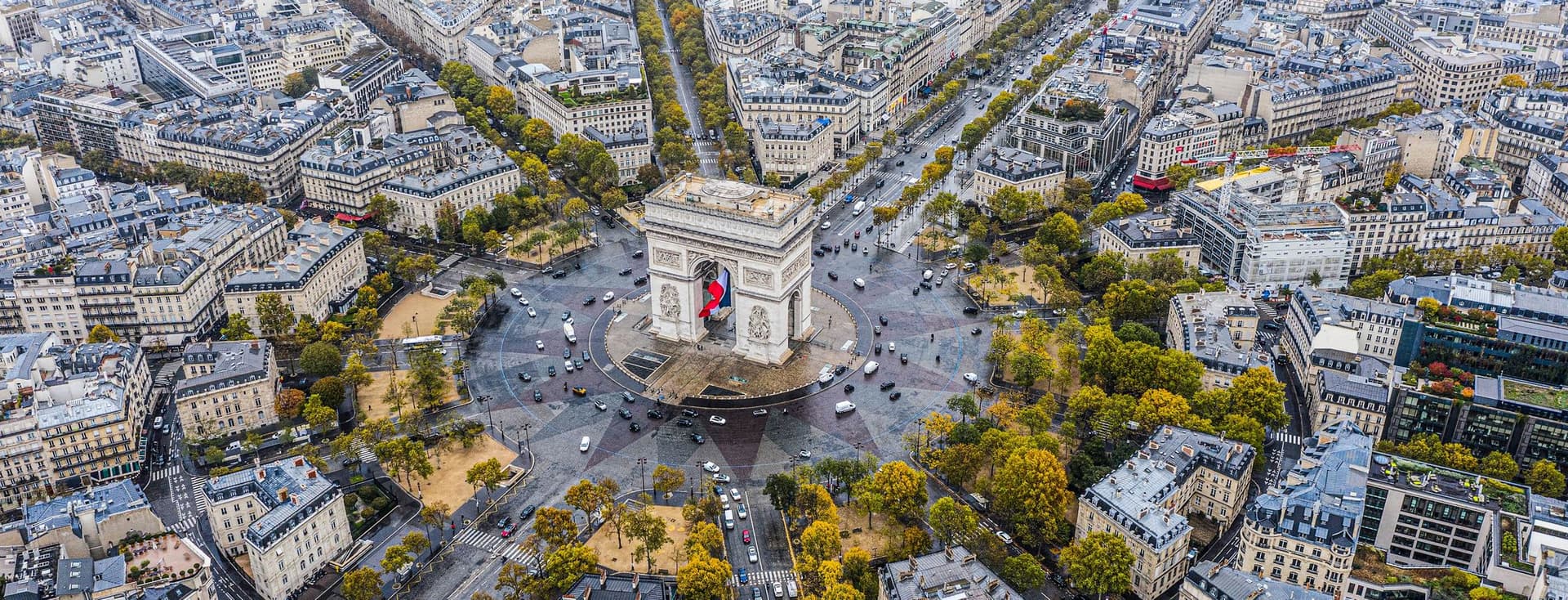 Aerial view of Paris, France, featuring the Arc de Triomphe at the center of a starburst of streets. Buildings and trees line the avenues.
