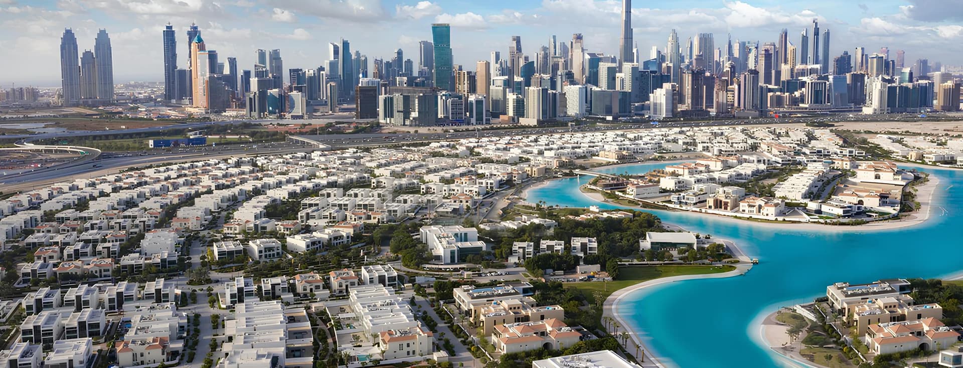 Aerial view of Dubai skyline with skyscrapers, residential areas, and a winding turquoise waterway under a partly cloudy sky.