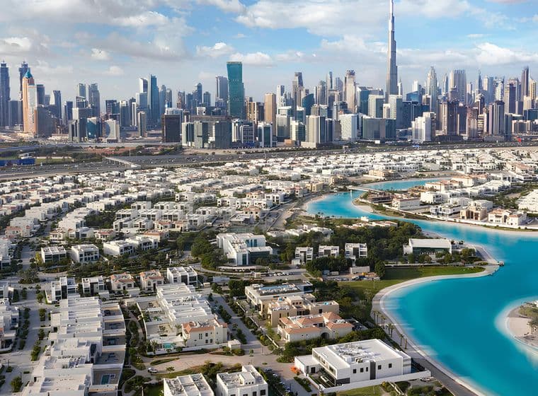 Aerial view of Dubai skyline with skyscrapers, residential areas, and a winding turquoise waterway under a partly cloudy sky.