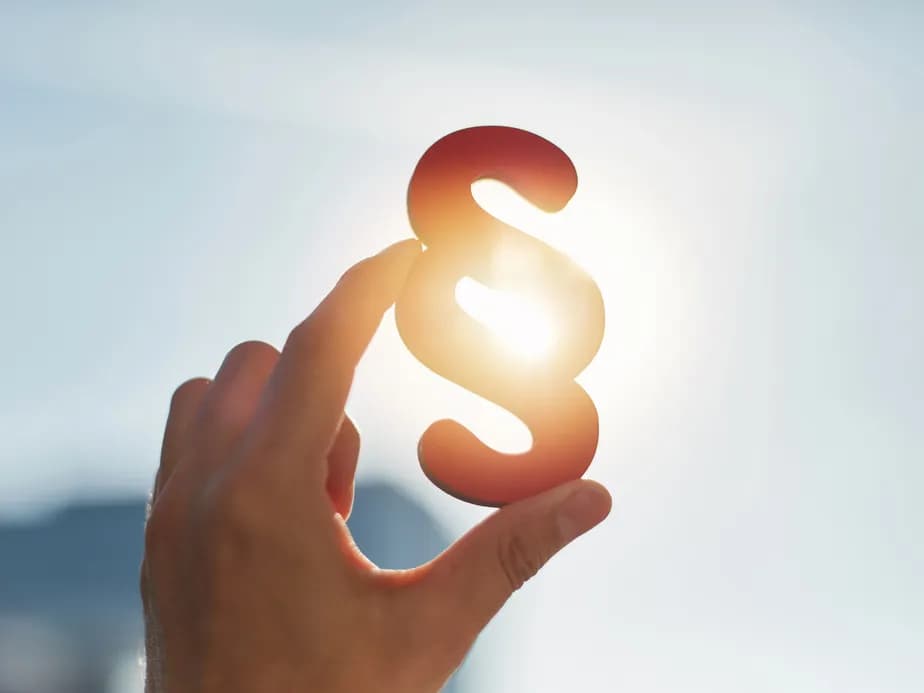 Hand silhouetted against a bright sky holding up a paragraph symbol with sun shining through, creating a backlit effect