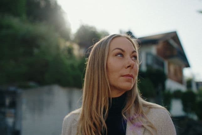 Portrait of a young woman with long blonde hair, looking upward, on a sunny suburban street with blurred trees and a house in the background.