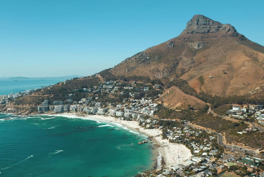 Aerial view of Camps Bay in Cape Town, South Africa. The beach with white sand and turquoise water stretches along the coastline. In the background rises the distinctive Lion's Head mountain. Residential areas and buildings extend between the beach and the mountainside. The image showcases the characteristic landscape of South Africa's Atlantic seaboard.