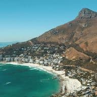 Aerial view of Camps Bay in Cape Town, South Africa. The beach with white sand and turquoise water stretches along the coastline. In the background rises the distinctive Lion's Head mountain. Residential areas and buildings extend between the beach and the mountainside. The image showcases the characteristic landscape of South Africa's Atlantic seaboard.