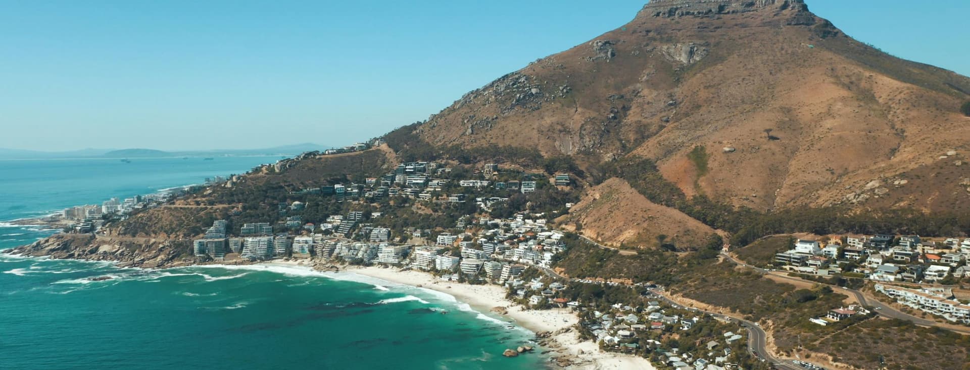 Aerial view of Camps Bay in Cape Town, South Africa. The beach with white sand and turquoise water stretches along the coastline. In the background rises the distinctive Lion's Head mountain. Residential areas and buildings extend between the beach and the mountainside. The image showcases the characteristic landscape of South Africa's Atlantic seaboard.