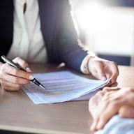 Close-up of two people in business attire reviewing a document with highlighted text on a wooden desk, with a notebook and glasses visible in the frame