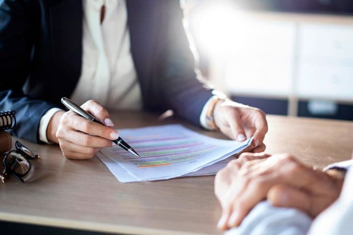 Close-up of two people in business attire reviewing a document with highlighted text on a wooden desk, with a notebook and glasses visible in the frame