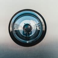 Close-up view of a circular security camera lens with a black outer ring and reflective glass surface mounted on a metallic wall. The camera's fisheye lens creates a distinctive circular reflection pattern.