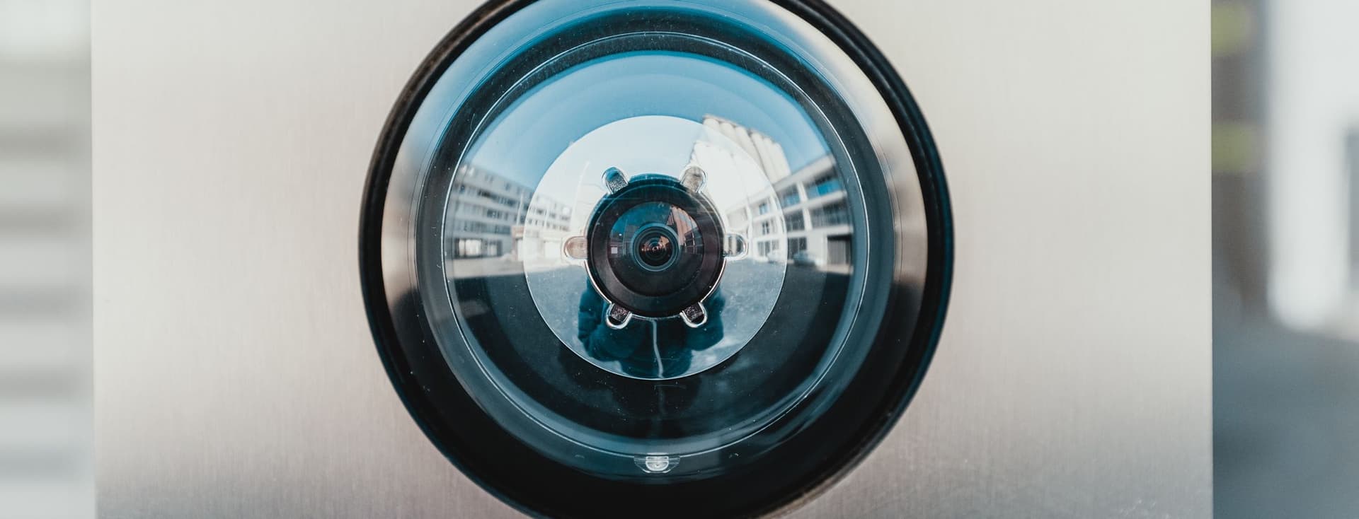Close-up view of a circular security camera lens with a black outer ring and reflective glass surface mounted on a metallic wall. The camera's fisheye lens creates a distinctive circular reflection pattern.