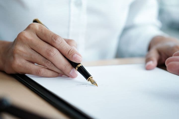 Une personne signe un document avec un stylo plume noir et doré sur un presse-papiers noir.