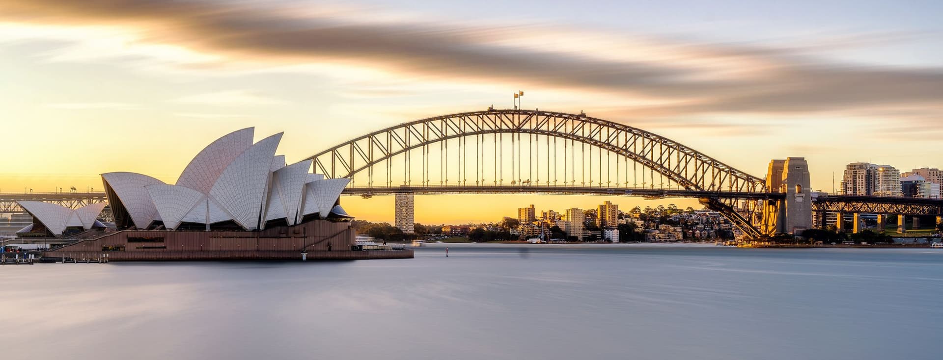 Sydney Opera House and Harbour Bridge at sunset. The water is smooth, and the sky is blue and orange.