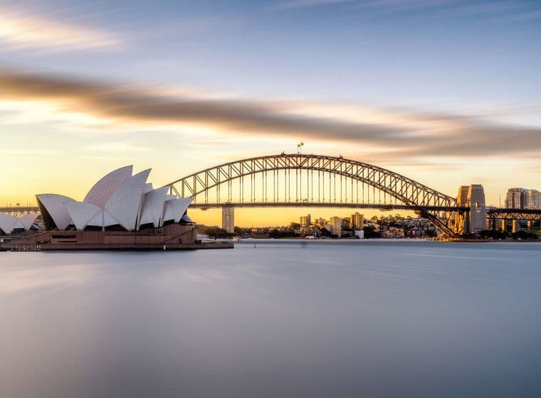 Sydney Opera House and Harbour Bridge at sunset. The water is smooth, and the sky is blue and orange.