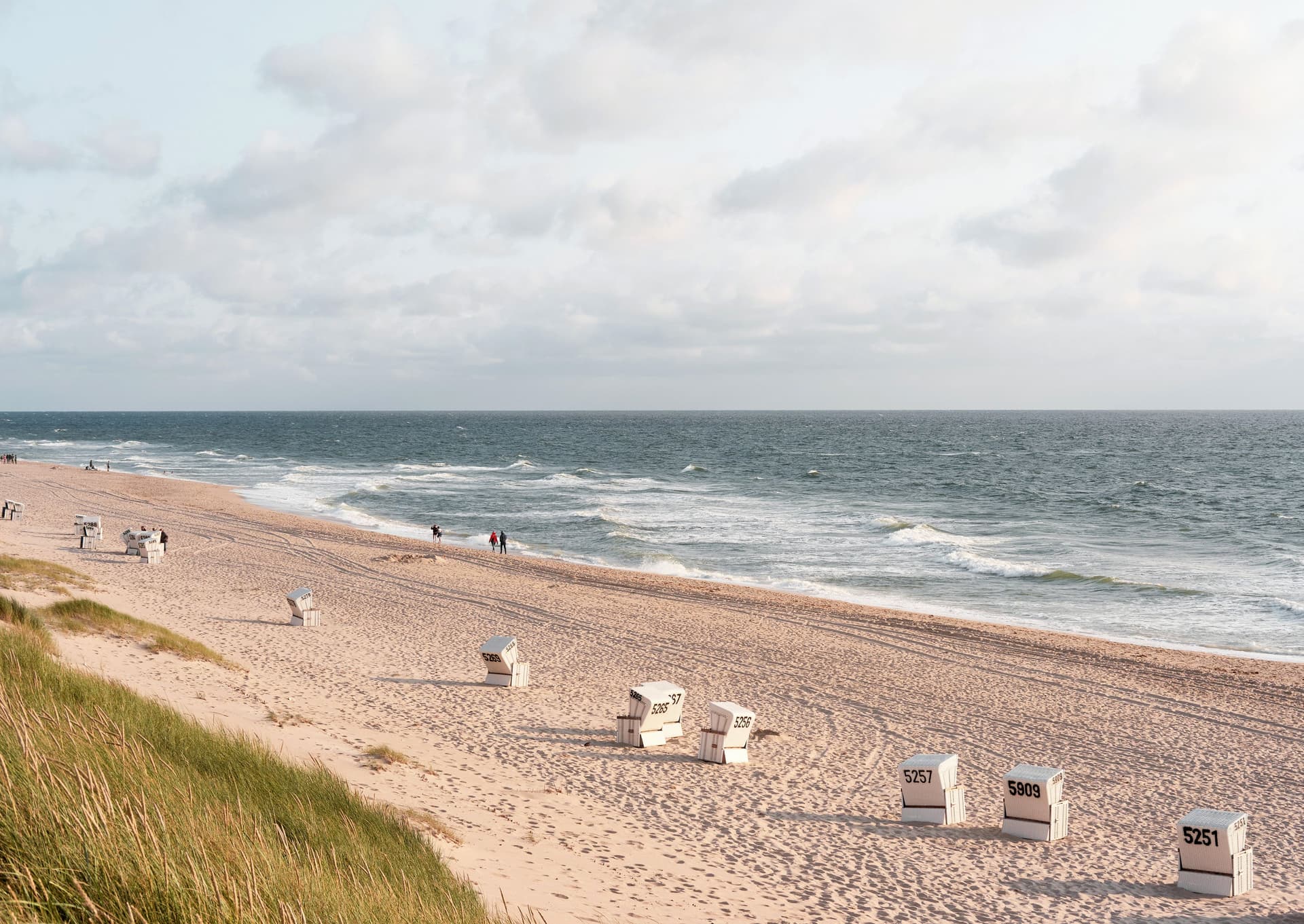 Beach scene with numbered white wicker chairs on sand, ocean waves, and cloudy sky.