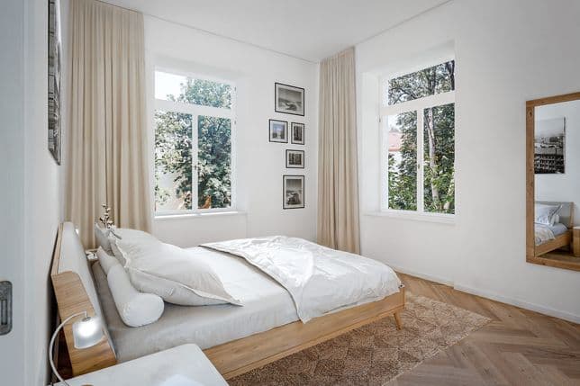 Bedroom featuring a wooden bed with white bedding and a light upholstered headboard. The room has herringbone wooden flooring, a beige area rug, and two tall windows with long light-colored curtains overlooking mature trees. A gallery of black-and-white photographs hangs between the windows, and a full-length mirror with a wooden frame stands on the right wall. Obrázek vygenerovaný umělou inteligencí