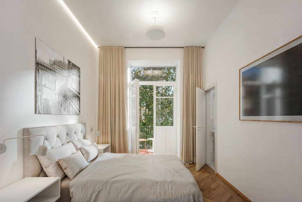 Bright bedroom with a double bed in neutral tones, complemented by bedside tables and wall-mounted lighting. Large glass doors lead to a balcony and bring in plenty of natural light, with curtains on the sides and artwork on the walls.