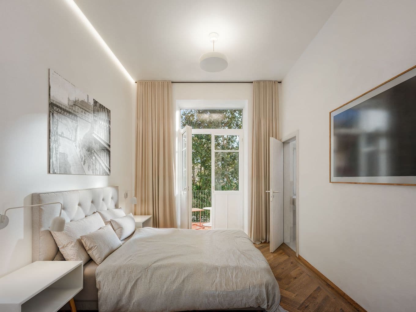 Bright bedroom with a double bed in neutral tones, complemented by bedside tables and wall-mounted lighting. Large glass doors lead to a balcony and bring in plenty of natural light, with curtains on the sides and artwork on the walls.