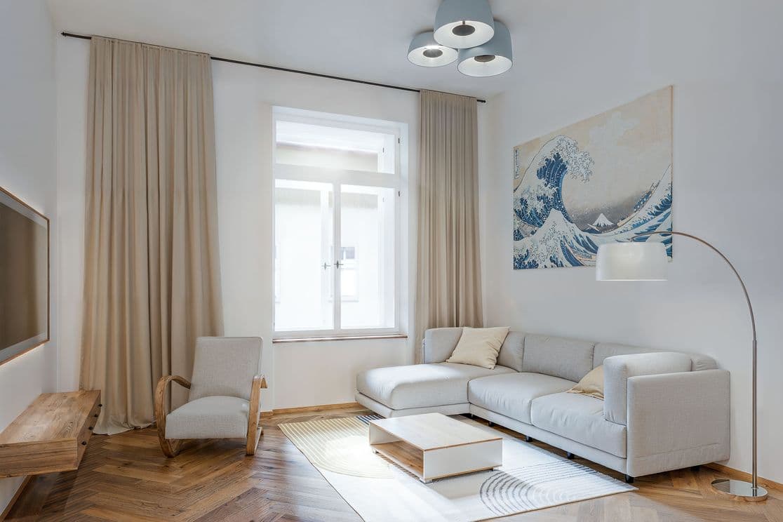 Bright open-plan space combining a living room, dining area, and kitchen. In the foreground there is a sofa and a coffee table on a rug, followed by a dining table with chairs, and a kitchen with white cabinets and a blue backsplash. The wall is complemented by a built-in bookshelf and artwork.