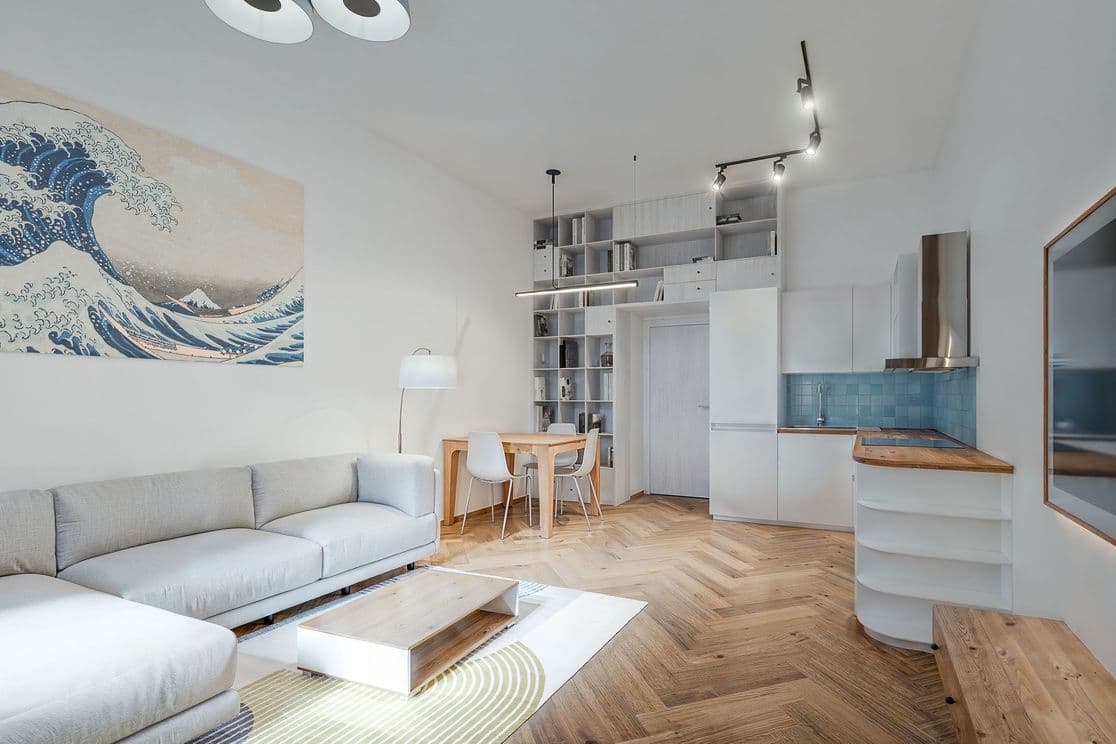 Bright open-plan space combining a living room, dining area, and kitchen. In the foreground there is a sofa and a coffee table on a rug, followed by a dining table with chairs, and a kitchen with white cabinets and a blue backsplash. The wall is complemented by a built-in bookshelf and artwork.