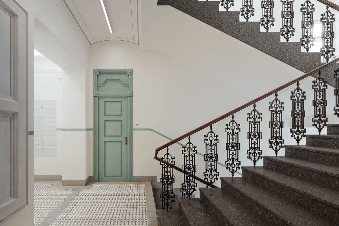 Bright entrance hall featuring a stone staircase with ornate metal railing and wooden handrail. The floor has patterned tiles, and in the background there is a white door and a corridor leading to other parts of the building.