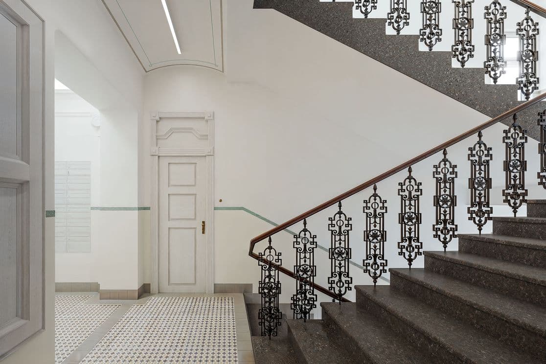 Bright entrance hall featuring a stone staircase with ornate metal railing and wooden handrail. The floor has patterned tiles, and in the background there is a white door and a corridor leading to other parts of the building.