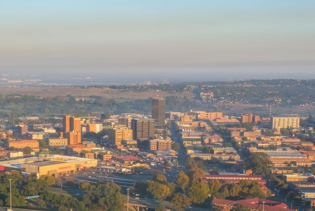 Aerial view of Johannesburg city skyline at dusk or dawn. The urban landscape shows a mix of high-rise buildings, commercial structures, and residential areas spreading into the distance. The foreground features some green spaces and trees, while the horizon shows a slight haze over the sprawling metropolitan area. This panoramic view captures South Africa's largest city with its distinctive urban development pattern.
