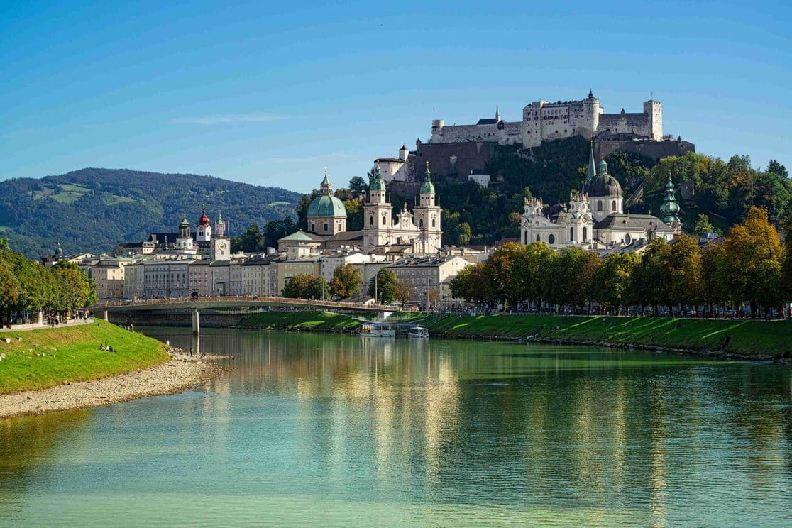 Salzburg Stadtansicht entlang der Salzach mit Festung Hohensalzburg, barocken Kirchenkuppeln und historischer Altstadt, gespiegelt im ruhigen Flusswasser.