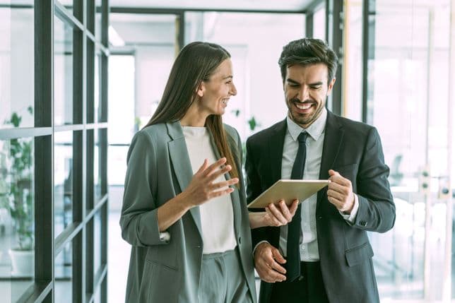 A smiling man and woman in business suits look at a tablet in a modern office.