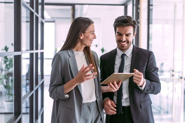 Dues persones de negocis somrients, una dona amb un vestit gris i un home amb un vestit negre, miren una tauleta en una oficina moderna.