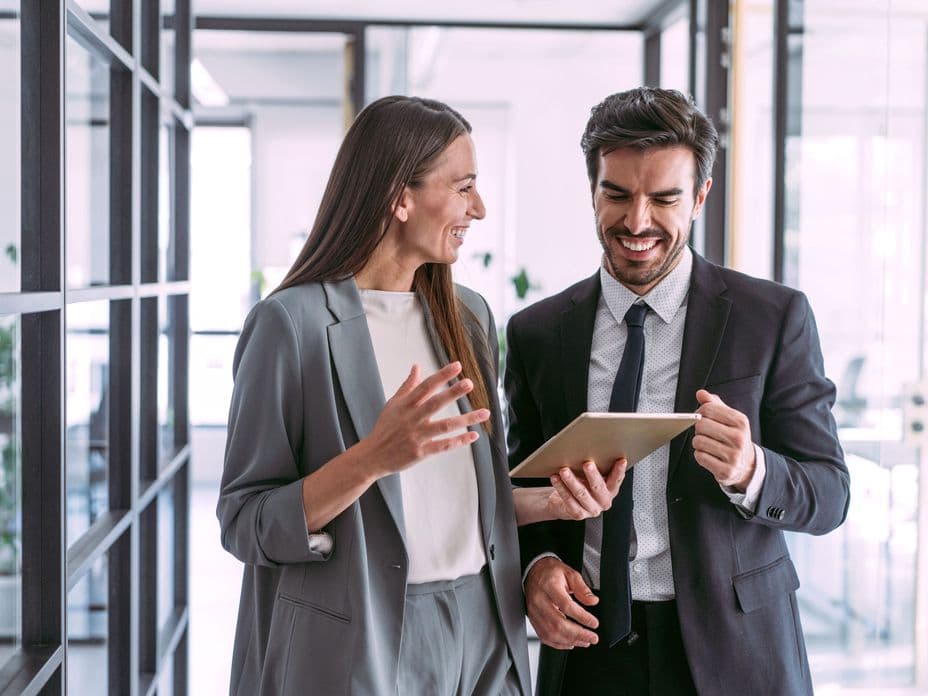 Zwei lächelnde Geschäftsleute, eine Frau in einem grauen Kostüm und ein Mann in einem schwarzen Anzug, schauen in einem modernen Büro auf ein Tablet.