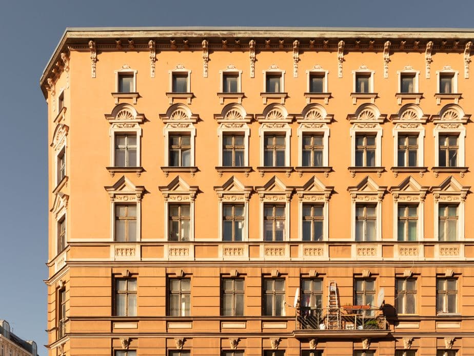 Ornate, sunlit, multi-story beige building with numerous windows and a small balcony, set against a clear blue sky.