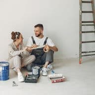 A couple sits on the floor with paint supplies, smiling at each other. A ladder leans against the white wall.
