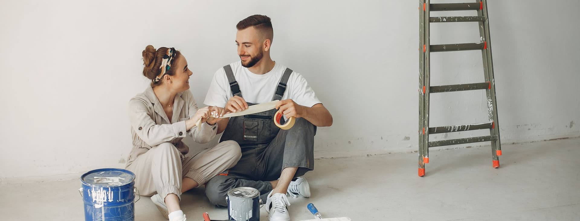 A couple sits on the floor with paint supplies, smiling at each other. A ladder leans against the white wall.