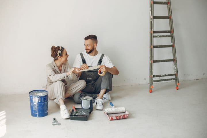 A couple sits on the floor with paint supplies, smiling at each other. A ladder leans against the white wall.