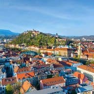 Aerial view of Graz with red-roofed buildings, a hilltop castle, and distant mountains under a clear blue sky.