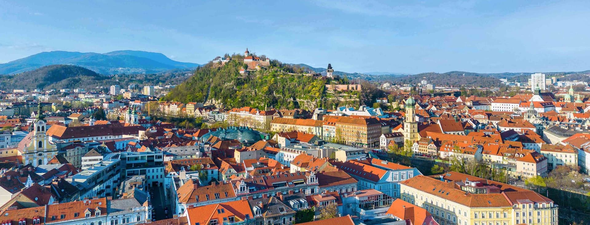 Luftaufnahme von Graz mit rotgedeckten Gebäuden, einer Burg auf einem Hügel und entfernten Bergen unter einem klaren blauen Himmel.