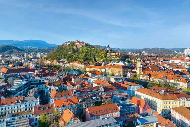 Aerial view of Graz with red-roofed buildings, a hilltop castle, and distant mountains under a clear blue sky.